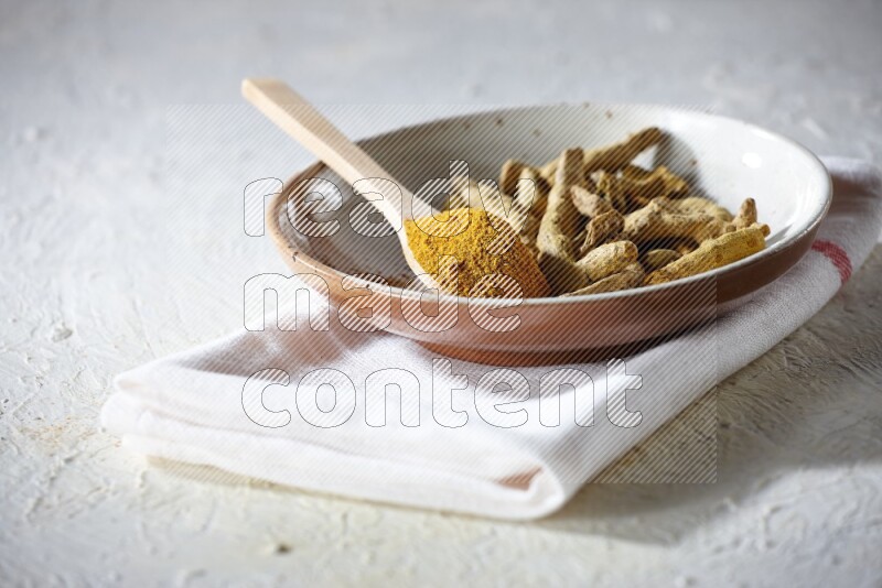 A plate filled with dried turmeric whole fingers and a wooden spoon full of turmeric powder on a textured white flooring