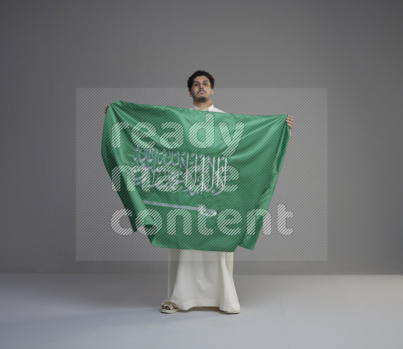 A Saudi man standing wearing thob holding big Saudi flag on gray background