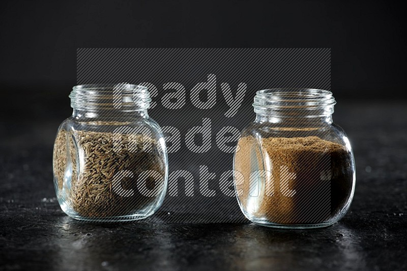 2 glass spice jars full of cumin powder and seeds on a textured black flooring