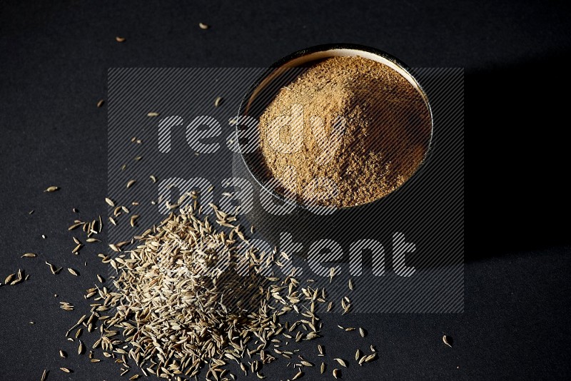A black pottery bowl full of cumin powder and spreaded cumin seeds on a black flooring