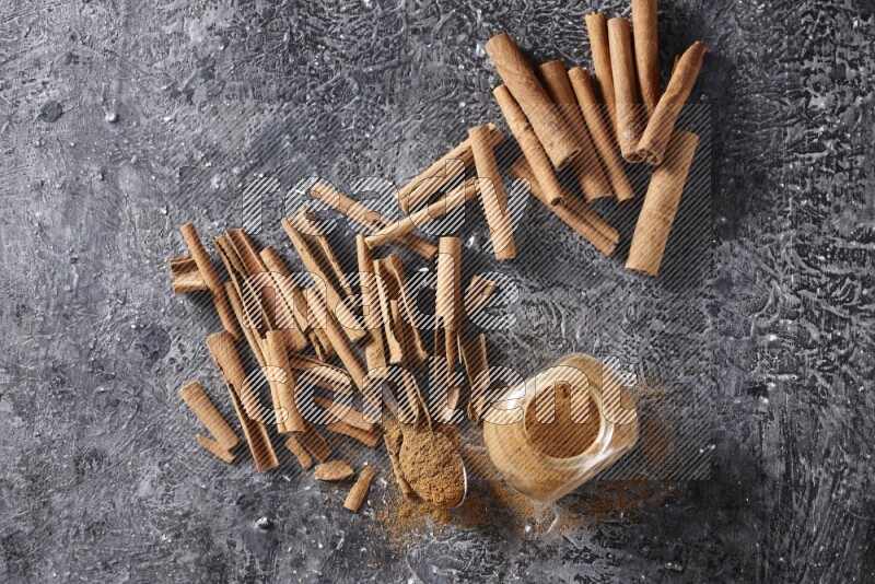 Herbal glass jar and a metal spoon full of cinnamon powder surrounded by cinnamon sticks on textured black background