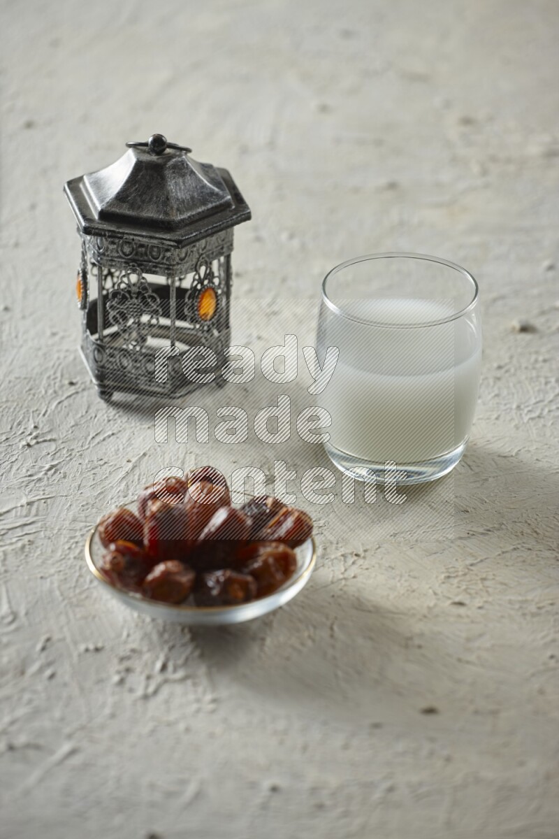 A silver lantern with different drinks, dates, nuts, prayer beads and quran on textured white background