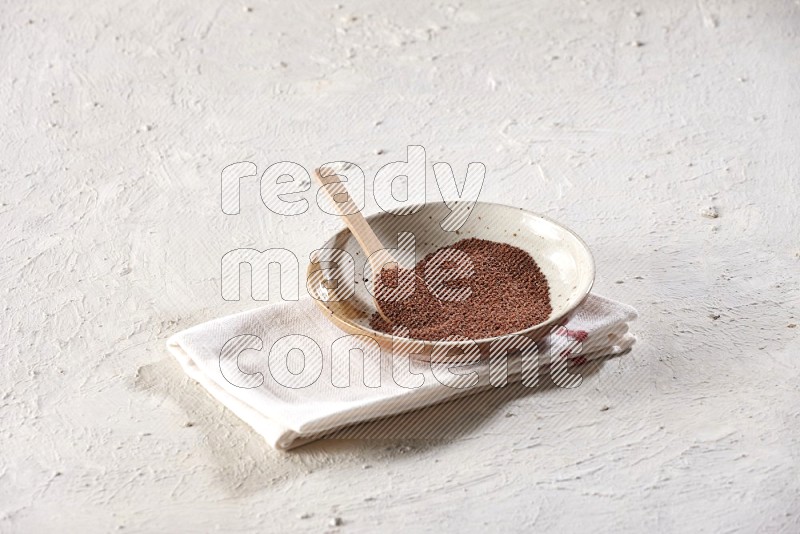 A multicolored pottery plate full of garden cress seeds with a wooden spoon full of the seeds on a napkin on a textured white flooring