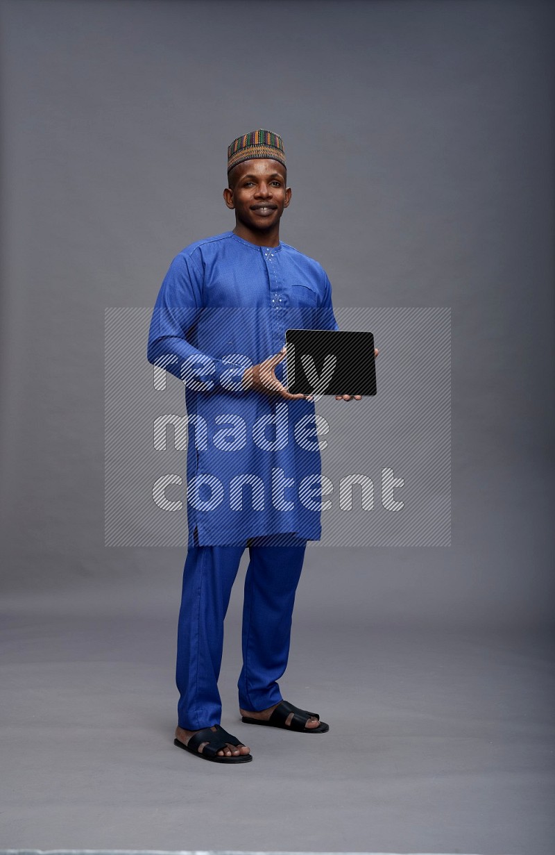 Man wearing Nigerian outfit standing showing tablet to camera on gray background