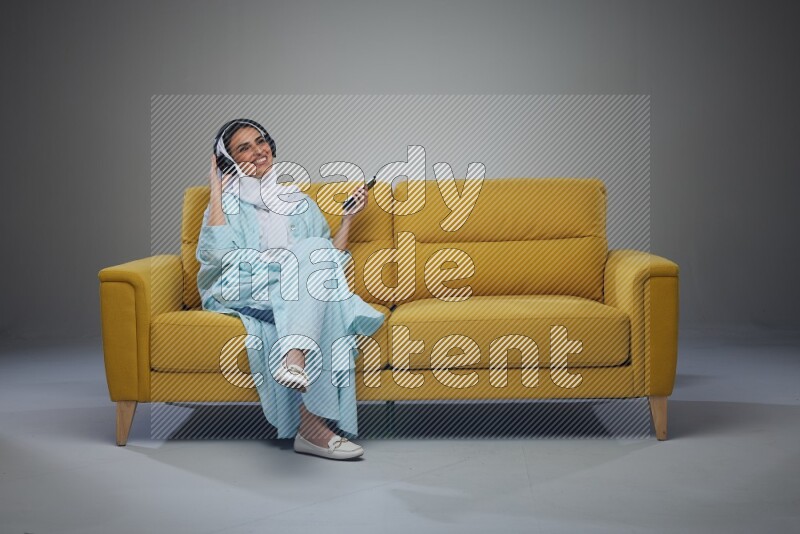 A Saudi woman wearing a light blue Abaya and a white head scarf sitting on a yellow sofa and holding an electronic while wearing headphone eye level on a grey background