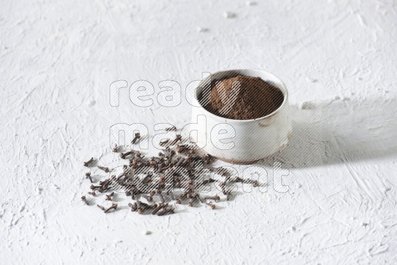 A beige ceramic bowl full of cloves powder and whole cloves on a white flooring