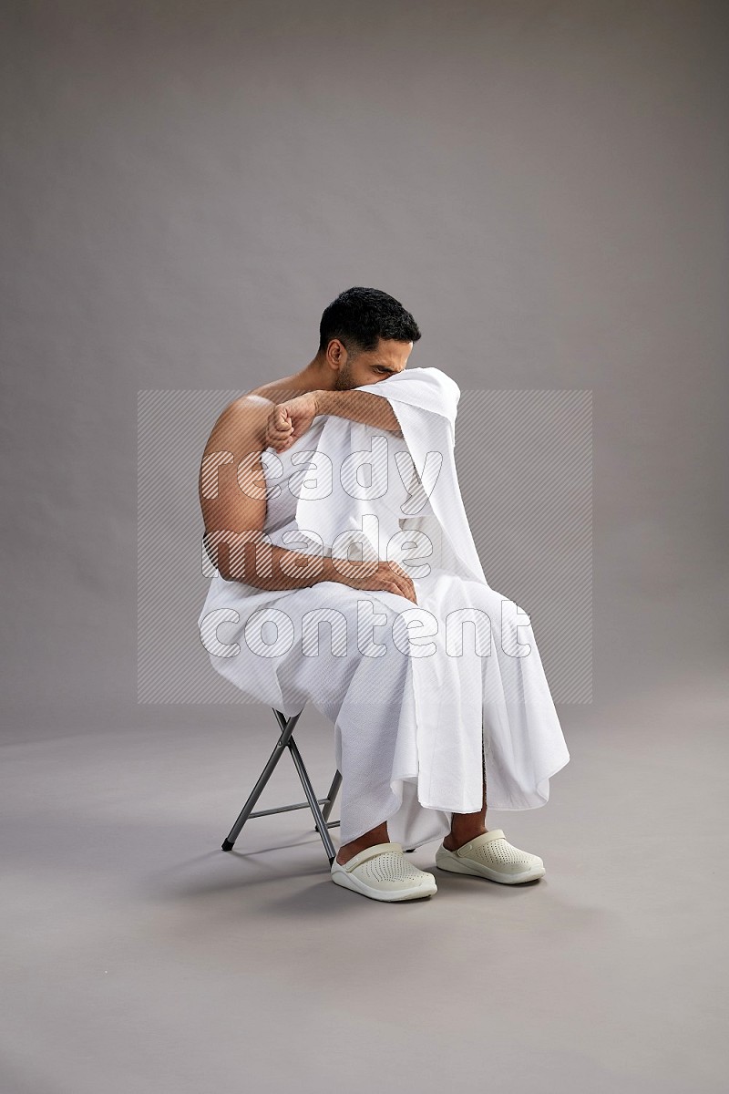 A man wearing Ehram sitting on chair showing how to sneeze on gray background