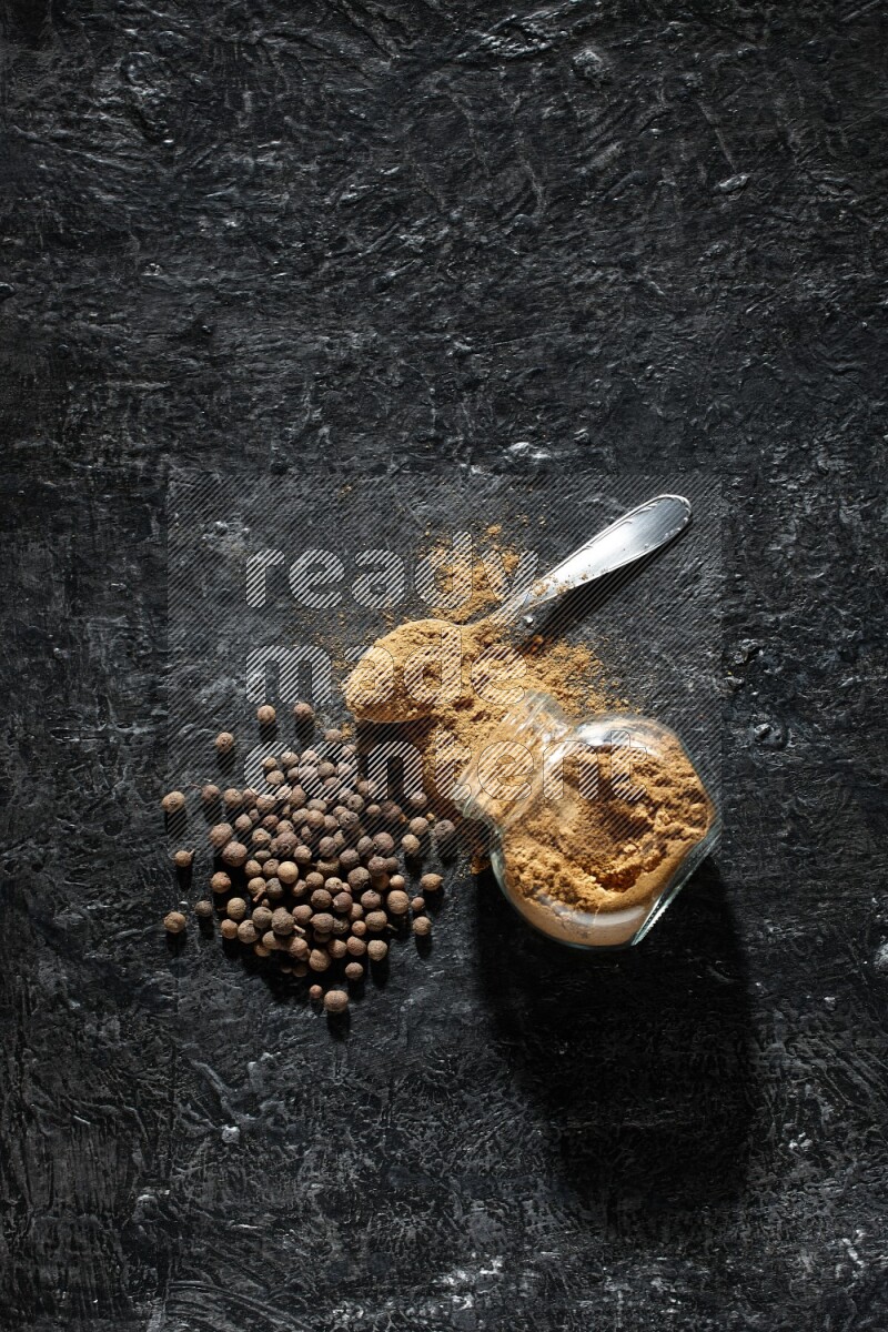 A flipped glass spice jar and metal spoon full of allspice powder and powder spilled out of it with whole balls on a textured black flooring