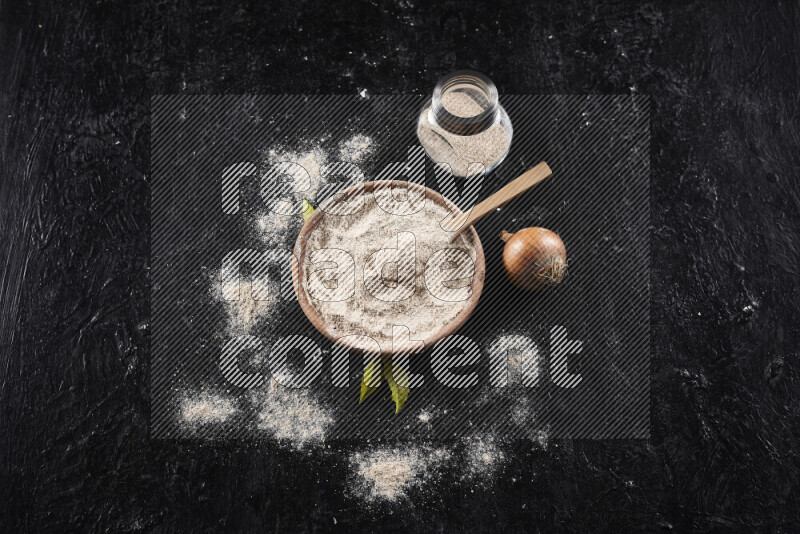 A wooden bowl full of onion powder with a glass jar beside it and fresh onion on black background