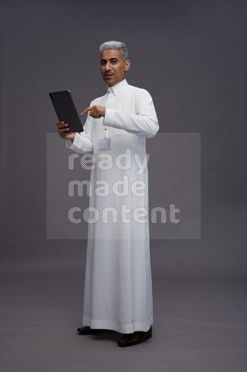 Saudi man wearing thob with neck strap employee badge standing working on tablet on gray background