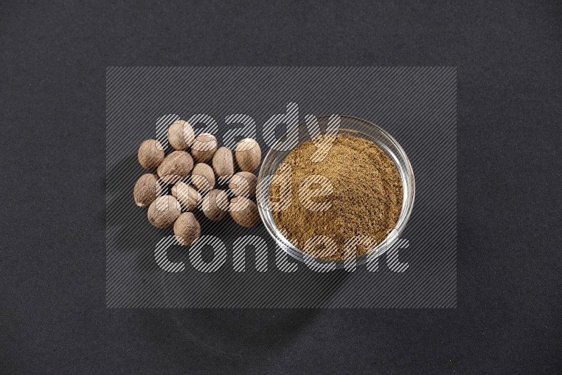 A glass bowl full nutmeg powder with the seeds beside it on a black flooring in different angles