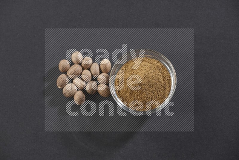 A glass bowl full nutmeg powder with whole seeds beside it on a black flooring
