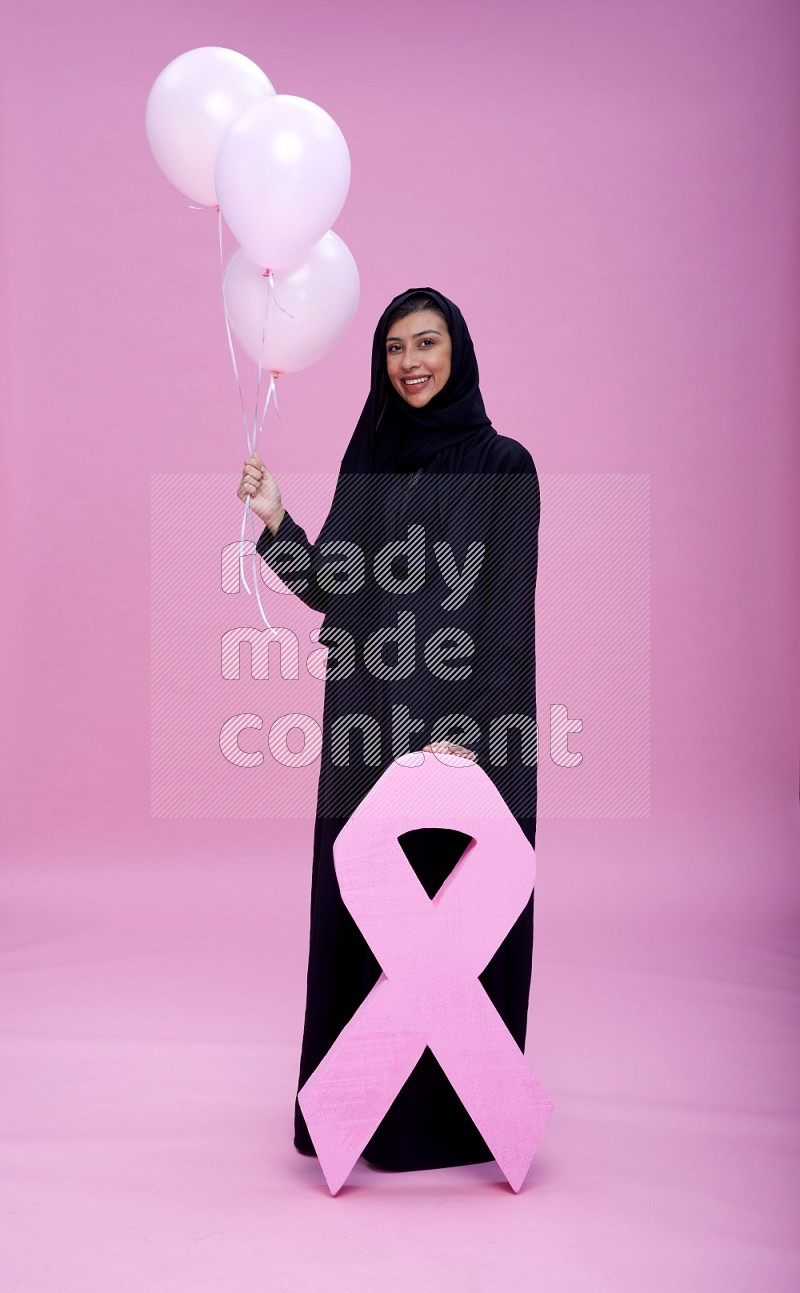 Saudi woman wearing pink ribbon on Abaya standing holding balloon with awareness on plain pink background