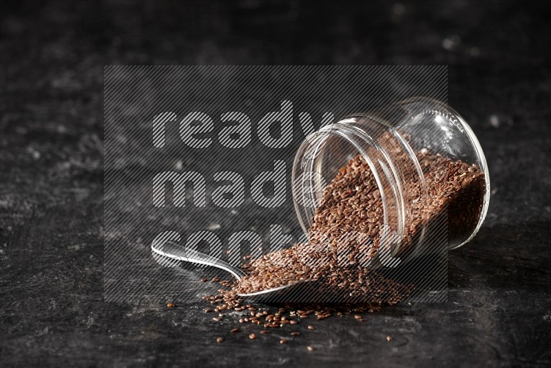 A glass jar full of flaxseeds flipped and seeds spread out with a metal spoon full of the seeds on a textured black flooring