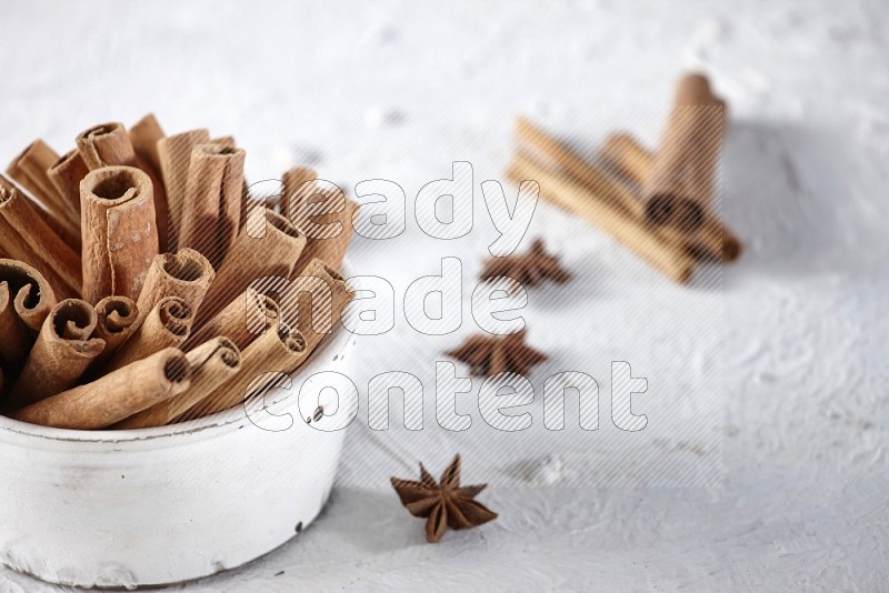 White bowl full of cinnamon sticks surrounded by star anis on a textured white background in different angles