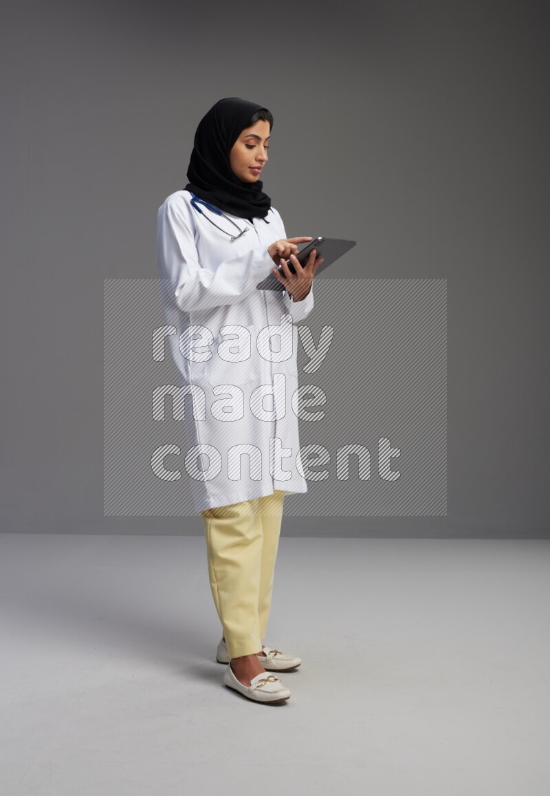 Saudi woman wearing lab coat with stethoscope standing working on tablet on Gray background