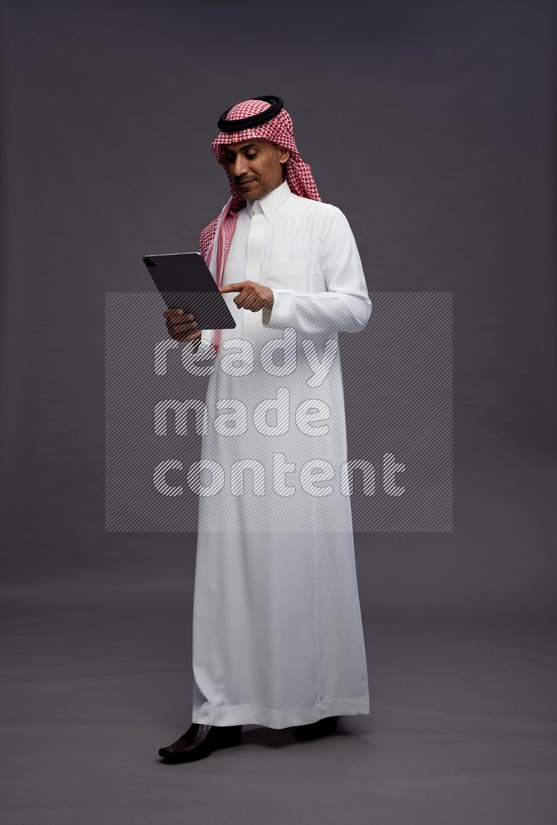 Saudi man wearing thob and shomag standing working on tablet on gray background