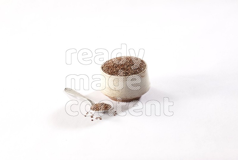 وعاء بيج فخاري ممتلئ بحبوب بذر الكتان مع ملعقه معدنية ممتلئة علي خلفية بيضاء