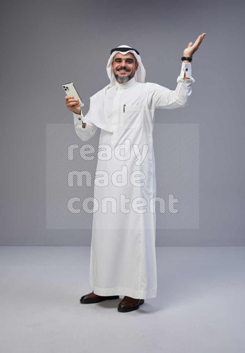 Saudi man Wearing Thob and white Shomag standing texting on phone on Gray background