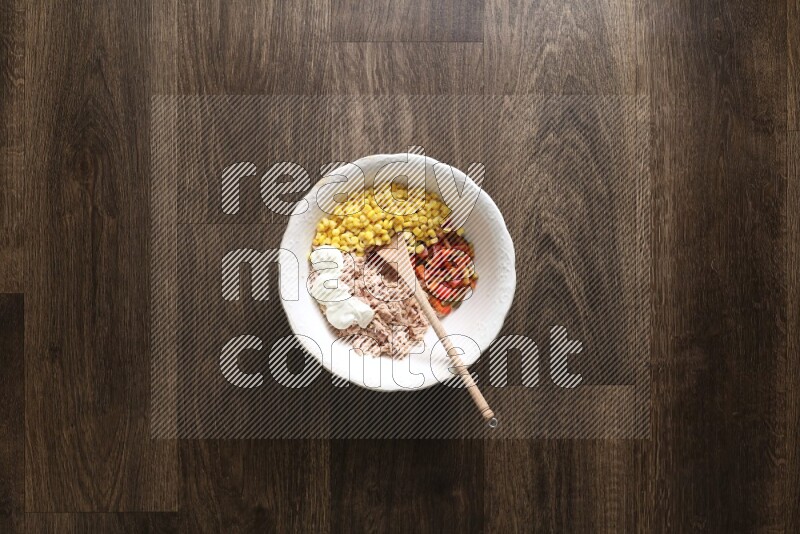 A white bowl full of tuna, colored bell pepper, sour cream, corn, parsley, black pepper powder and sauce, with wooden spoon on wooden background