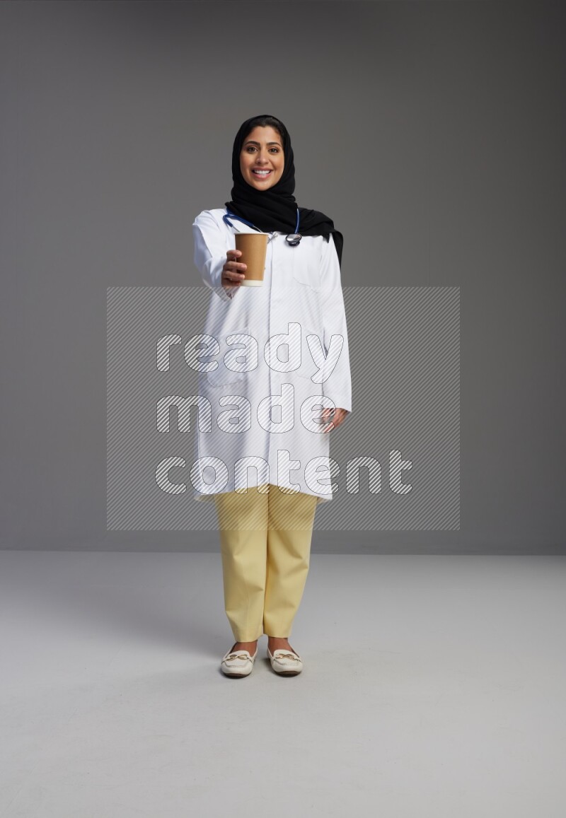 Saudi woman wearing lab coat with stethoscope standing holding paper cup on Gray background