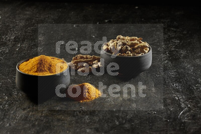 2 pottery black bowls, one full of turmeric powder and the other full of dried turmeric whole fingers on a textured black flooring