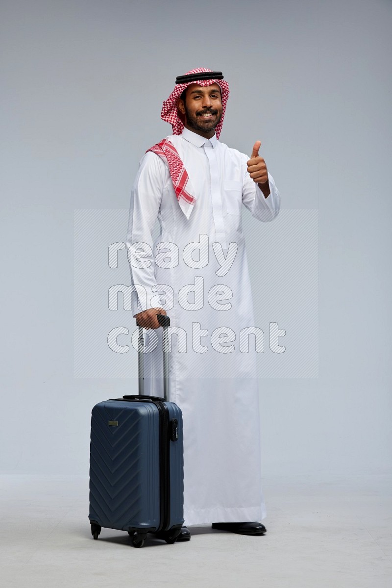 Saudi man wearing Thob and shomag standing holding Travel bag on Gray background