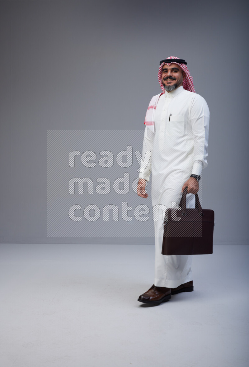 Saudi man Wearing Thob and red Shomag standing holding bag on Gray background