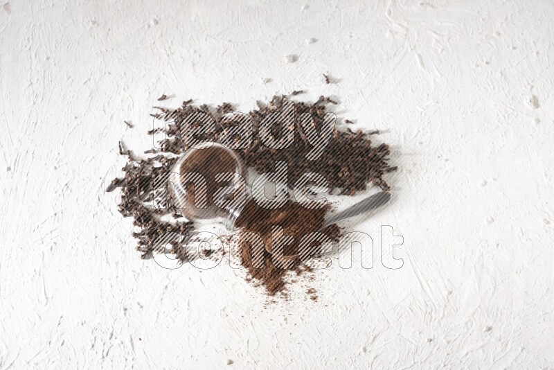 A flipped glass spice jar and a metal spoon full of cloves powder and powder came out of the jar with cloves spread on textured white flooring