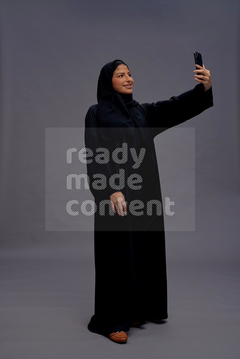 Saudi woman wearing Abaya standing taking selfie on gray background