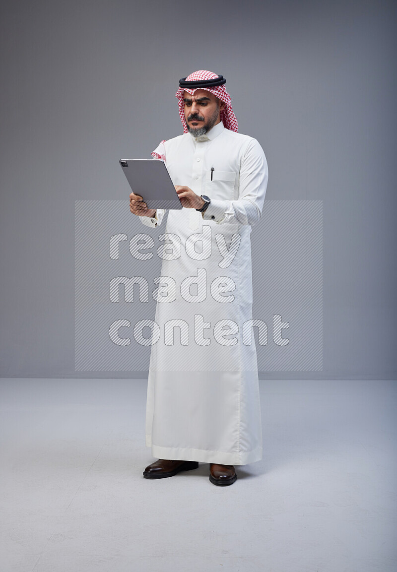 Saudi man Wearing Thob and red Shomag standing working on tablet on Gray background