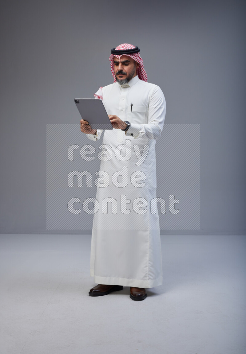 Saudi man Wearing Thob and red Shomag standing working on tablet on Gray background