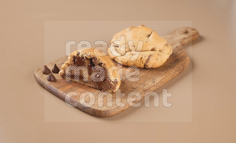 a chocolate chip cookie with another one cut in half on a wooden cutting board on a brown background