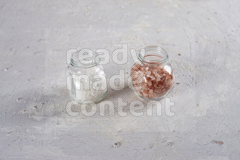 2 glass jars one is filled with coarse himalayan salt and the other with coarse sea salt on white background