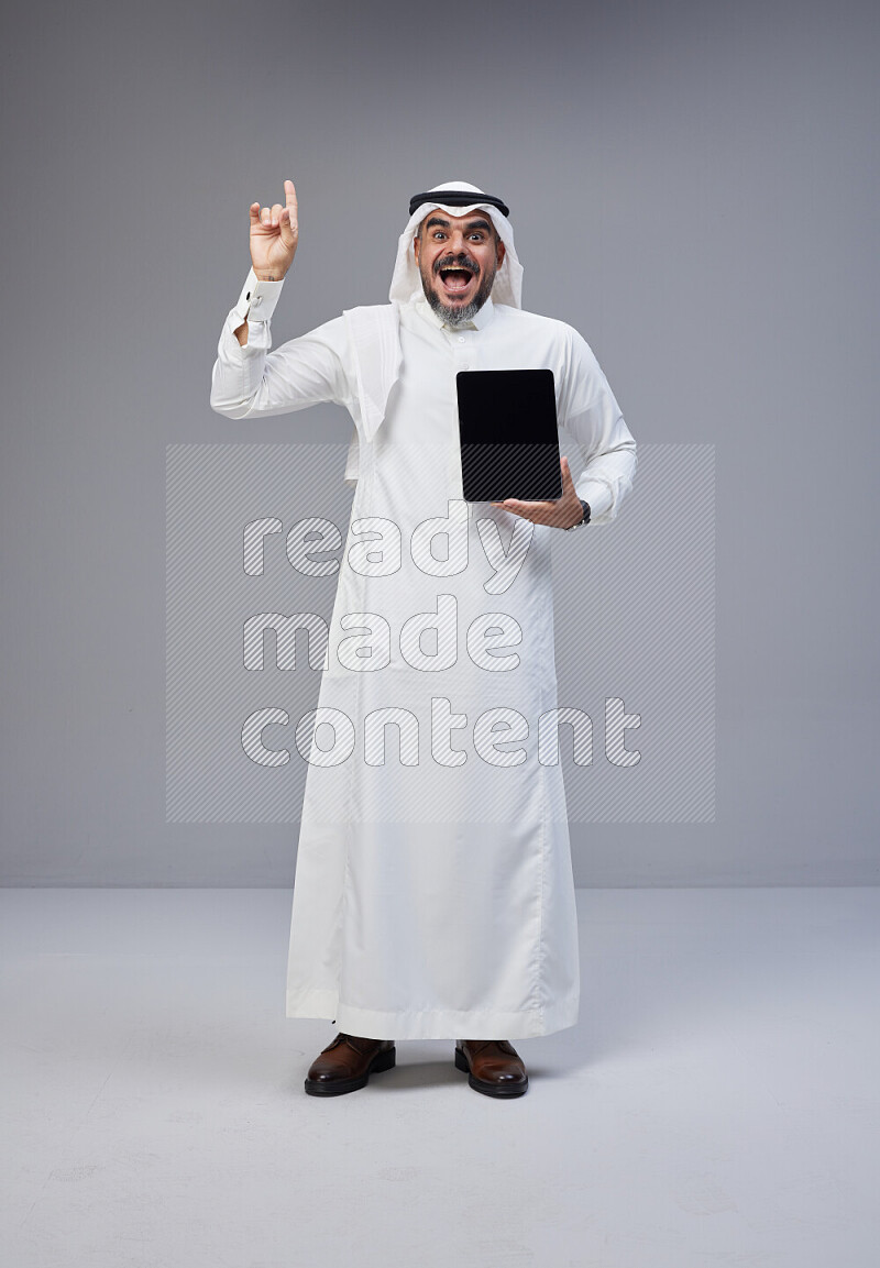 Saudi man Wearing Thob and white Shomag standing showing tablet to camera on Gray background