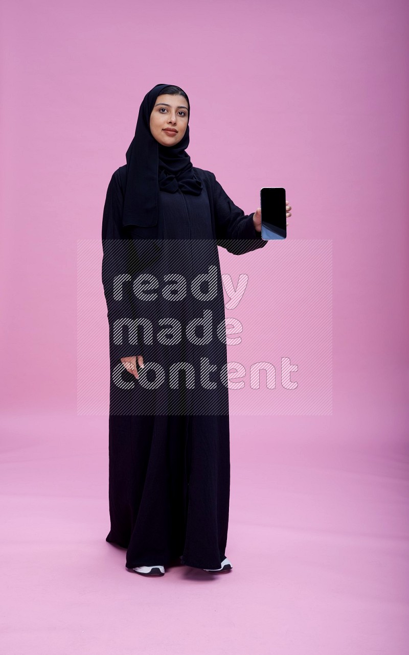 Saudi woman wearing Abaya standing showing phone to camera on pink background