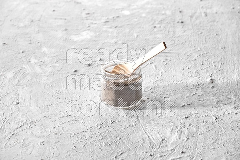 A glass jar full of black pepper powder with a wooden spoon on a textured white flooring