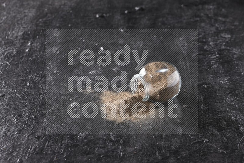 A flipped glass spice jar full of black pepper powder on textured black flooring
