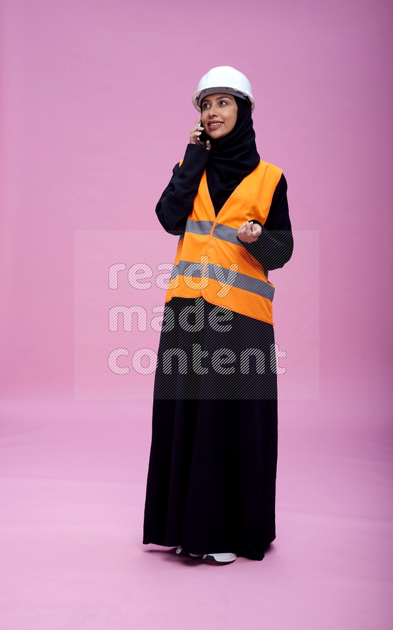 Saudi woman wearing Abaya with engineer vest and helmet standing talking on phone on pink background