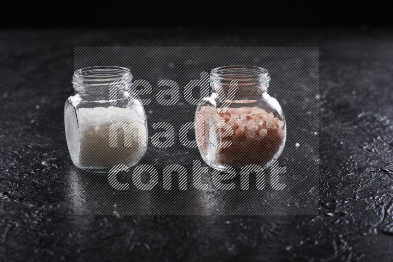 2 glass jars one is filled with coarse himalayan salt and the other with coarse sea salt on black background