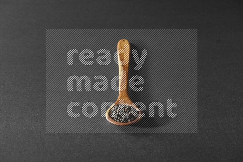 A wooden ladle full of black pepper beads on a black flooring