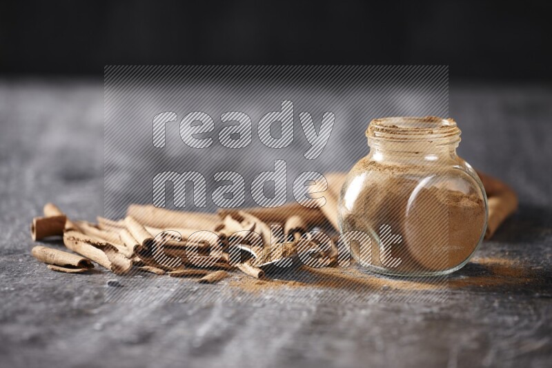 Herbal glass jar and a metal spoon full of cinnamon powder surrounded by cinnamon sticks on textured black background