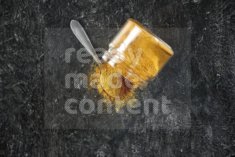A flipped glass jar and a metal spoon full of turmeric powder and powder spilled out of it on textured black flooring