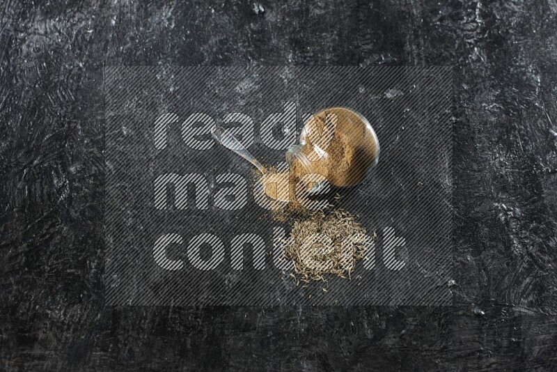 A flipped glass spice jar and a metal spoon full of cumin powder and powder spilled out with cumin seeds on a textured black flooring