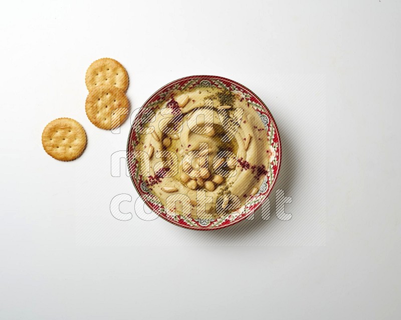 Hummus in a red plate with patterns garnished with zaatar & sumak on a white background