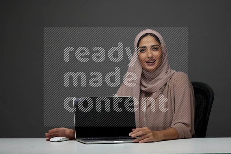 A woman Sitting on her desk  Pointing at her laptop on a Gray Background wearing Brown Abaya with Hijab