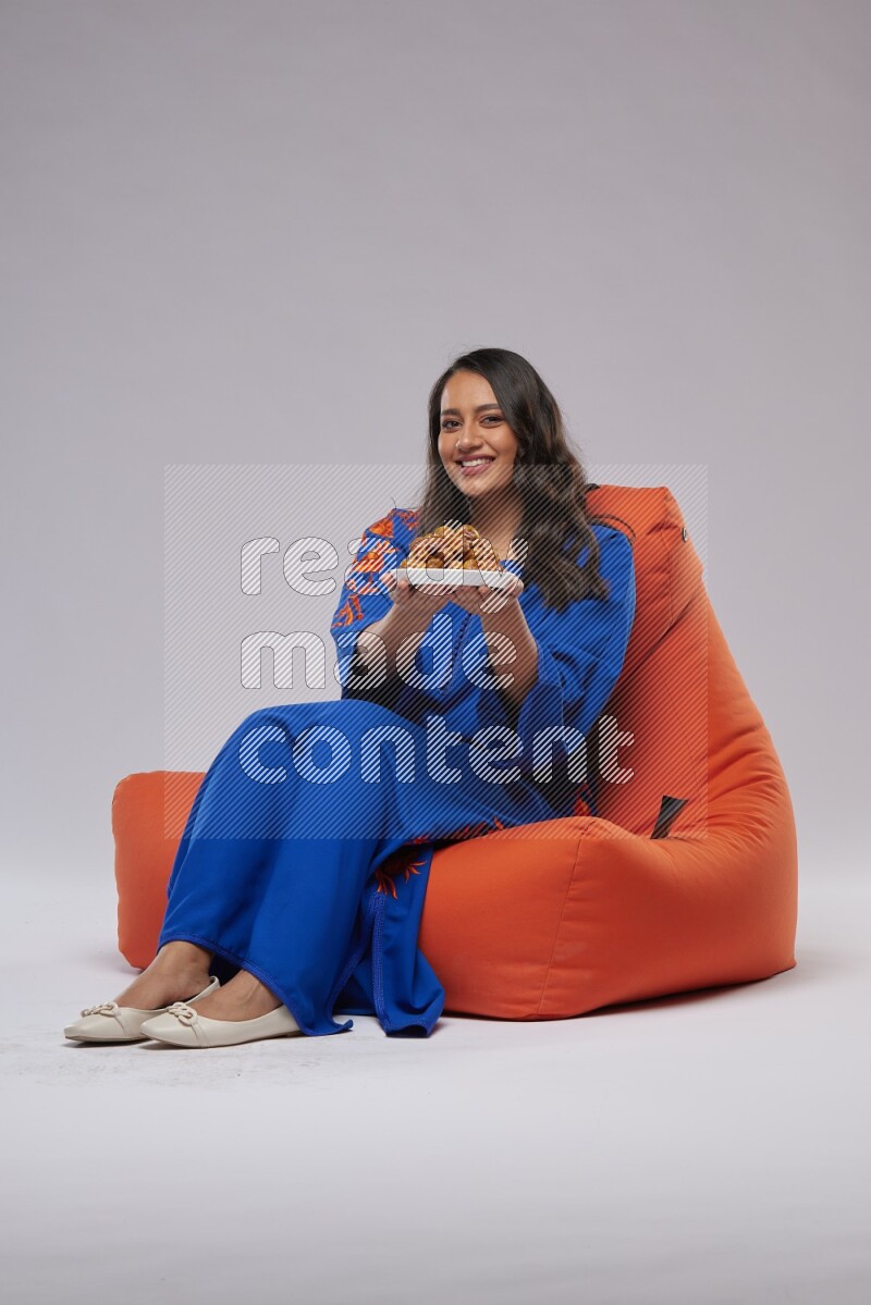 A Woman sitting on an orange beanbag wearing Jalabeya holding a plate of dates