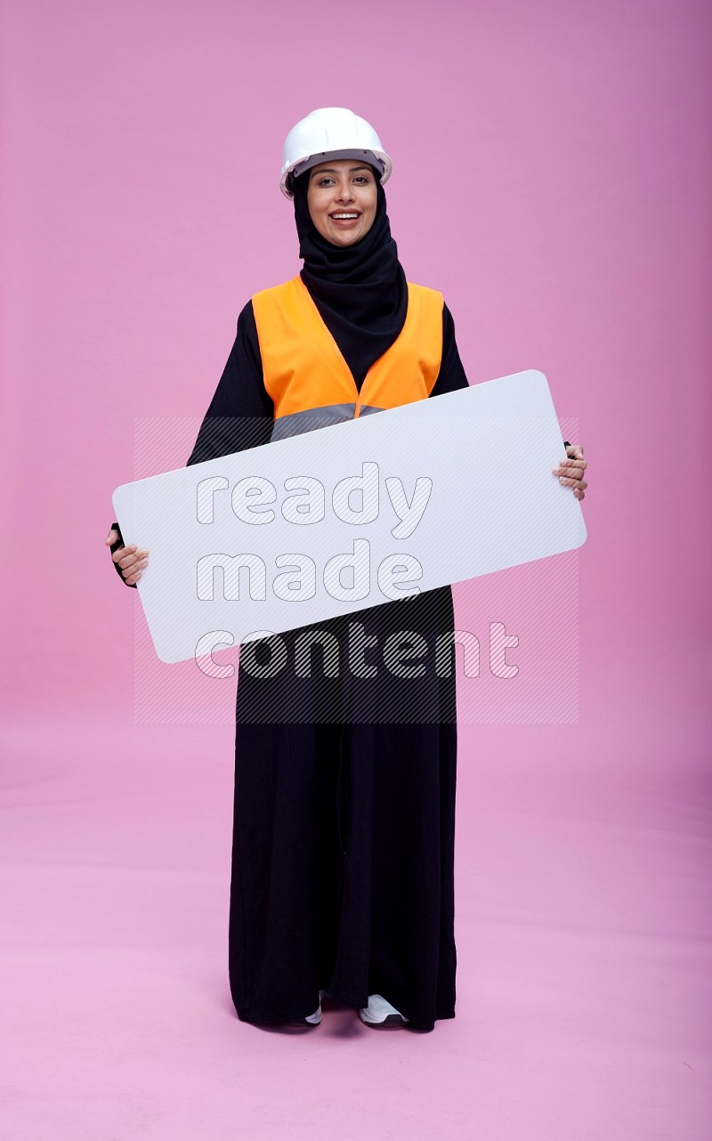 Saudi woman wearing Abaya with engineer vest and helmet standing holding board on pink background