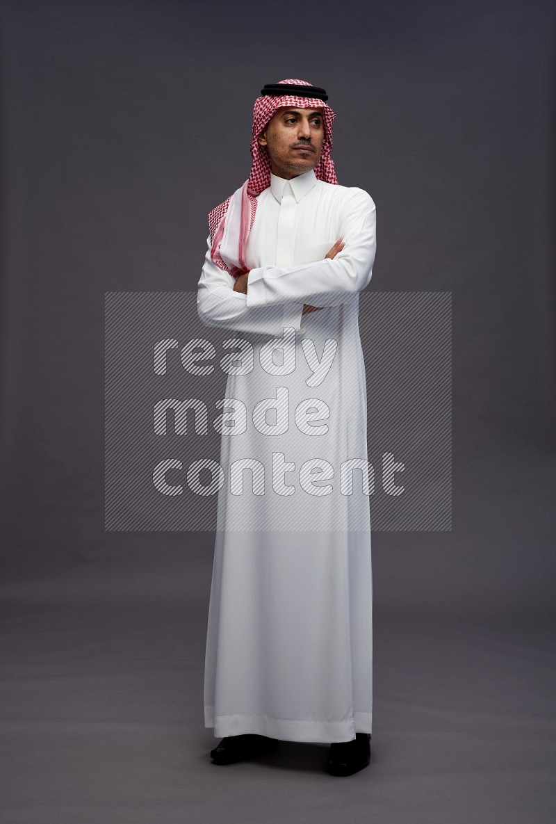 Saudi man wearing thob and shomag standing with crossed arms on gray background