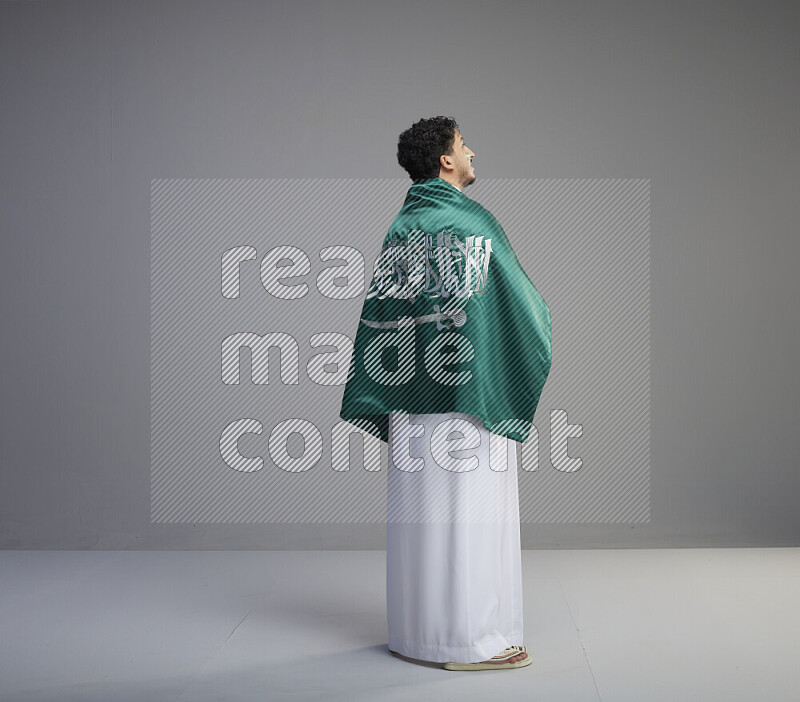 A Saudi man standing wearing thob with face painting wrapping big Saudi flag on gray background